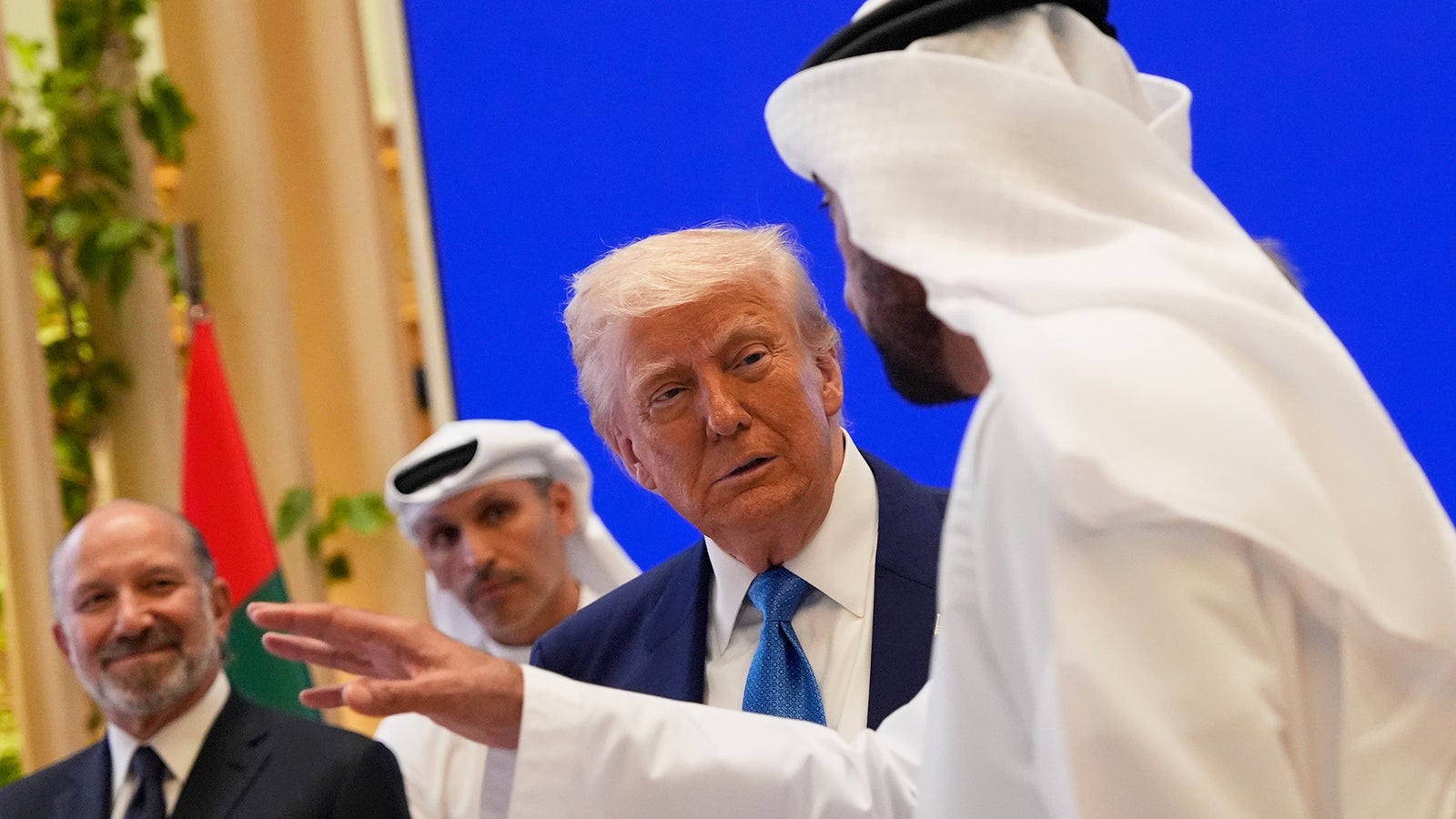 A group of three men are engaged in conversation, with one man gesturing, in front of a blue backdrop.