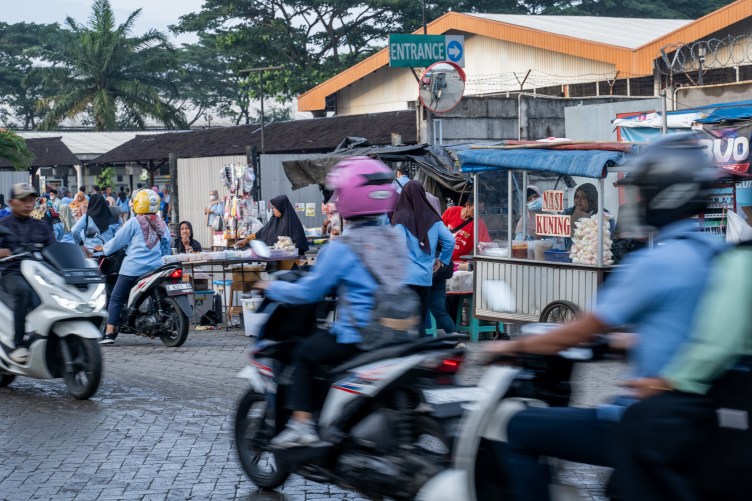 Numerous people on motorbikes and on foot, wearing blue uniforms, in front of a large building and a sign reading “Entrance.” Some people are buying items from vendors.