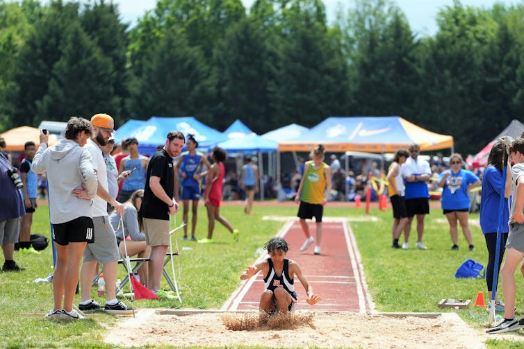 An outdoor sports events with a young woman landing a long jump in the foreground