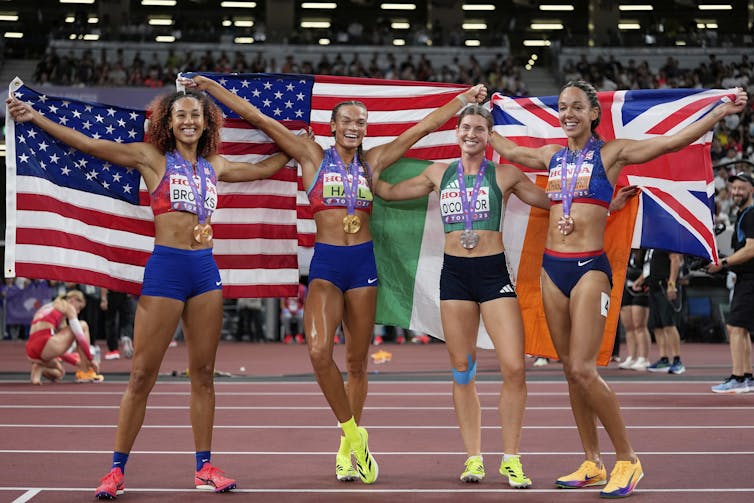 Four women wearing athletic clothes, draped in flags on an athletic track