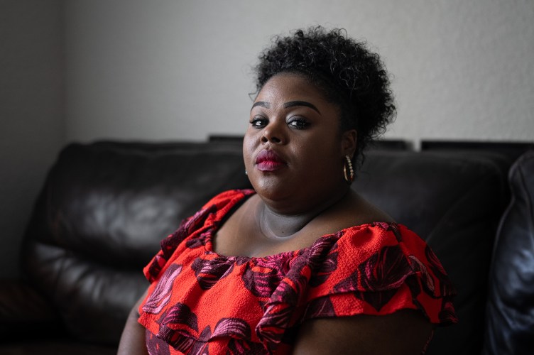 A woman looks directly at the camera wearing a red floral dress and earrings. She sits on a dark leather couch.