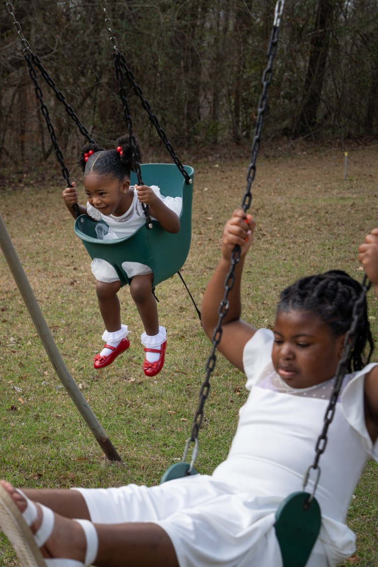 Two children wearing white dresses swing on a green swing set.