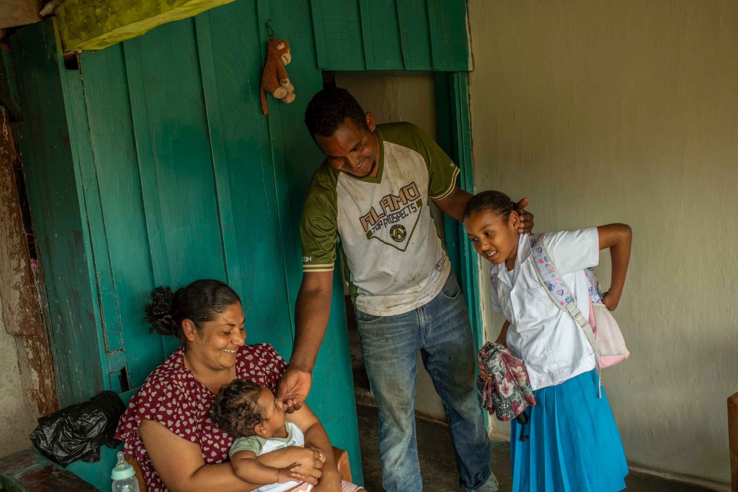 A woman wearing a red blouse with pink hearts holds an infant on her lap. A man wearing dirt-covered jeans touches the child’s face with one hand and holds the head of a young girl wearing a white shirt and blue skirt. They are in a house with an aqua wall in the background.