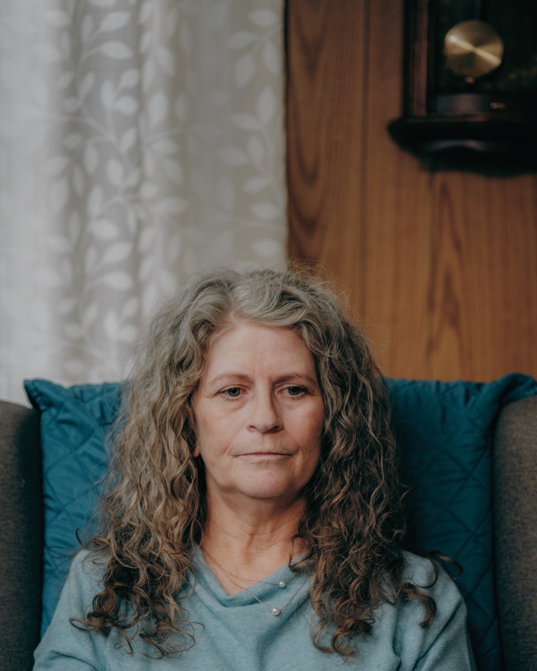 A woman with long hair, wearing a blue shirt, sits on a dark, leather sofa in a living room with wood-panneled walls.