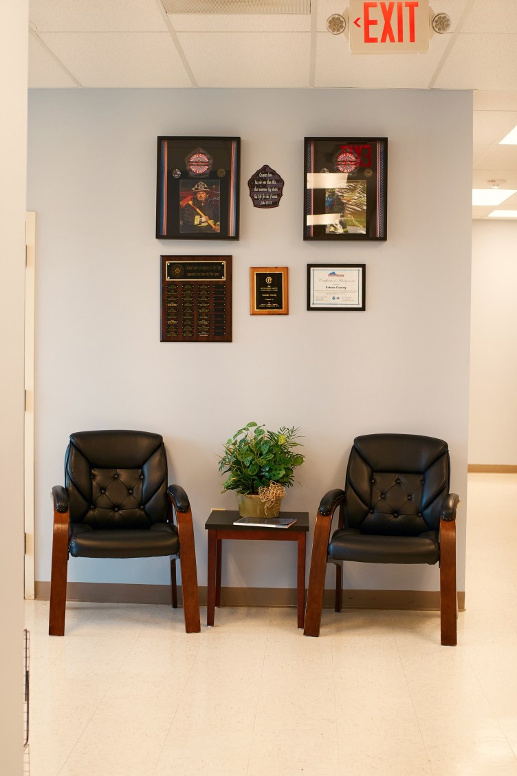 Two framed photos of men in firefighter uniforms hang on a wall with other framed pieces above two chairs and an end table.