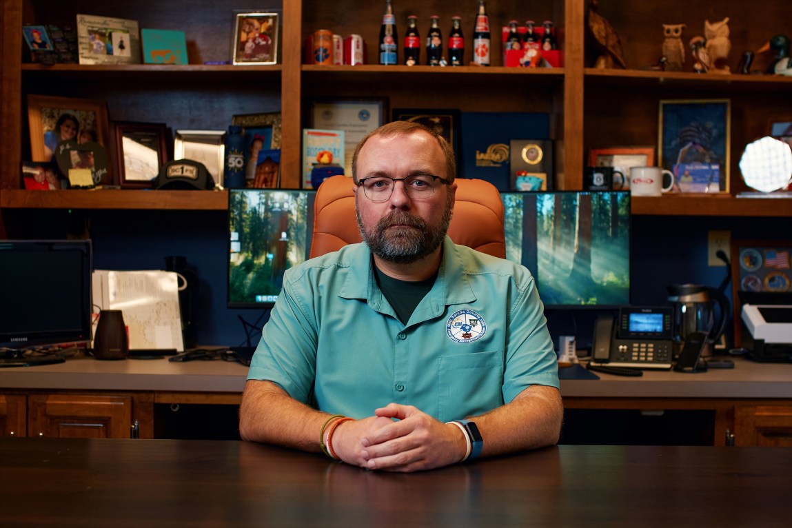 A man with glasses and a beard wears an aqua button-up shirt with an embroidered patch. He sits at a wooden desk in front of two computer monitors, a desk phone and various knickknacks.