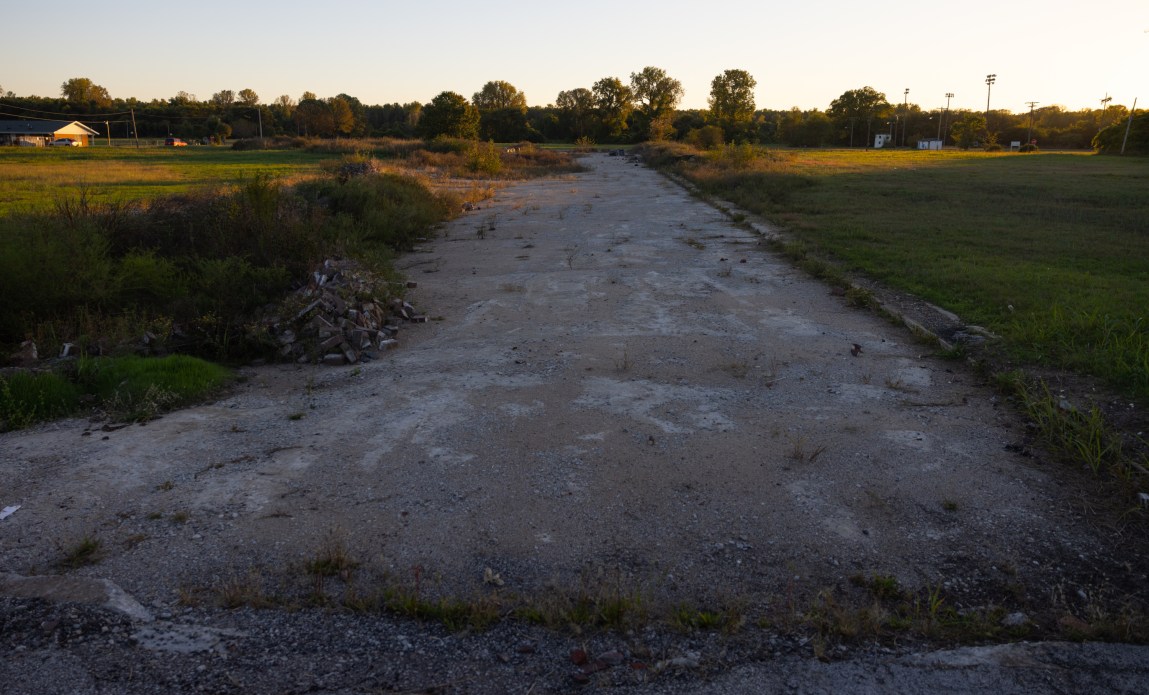 A patch of dirt and gravel sits vacant in the middle of a field, with houses in the background.