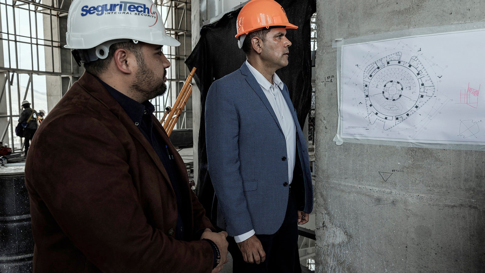 Two men, one wearing a hard hat and the other in a suit, examine architectural plans on a wall at a construction site.