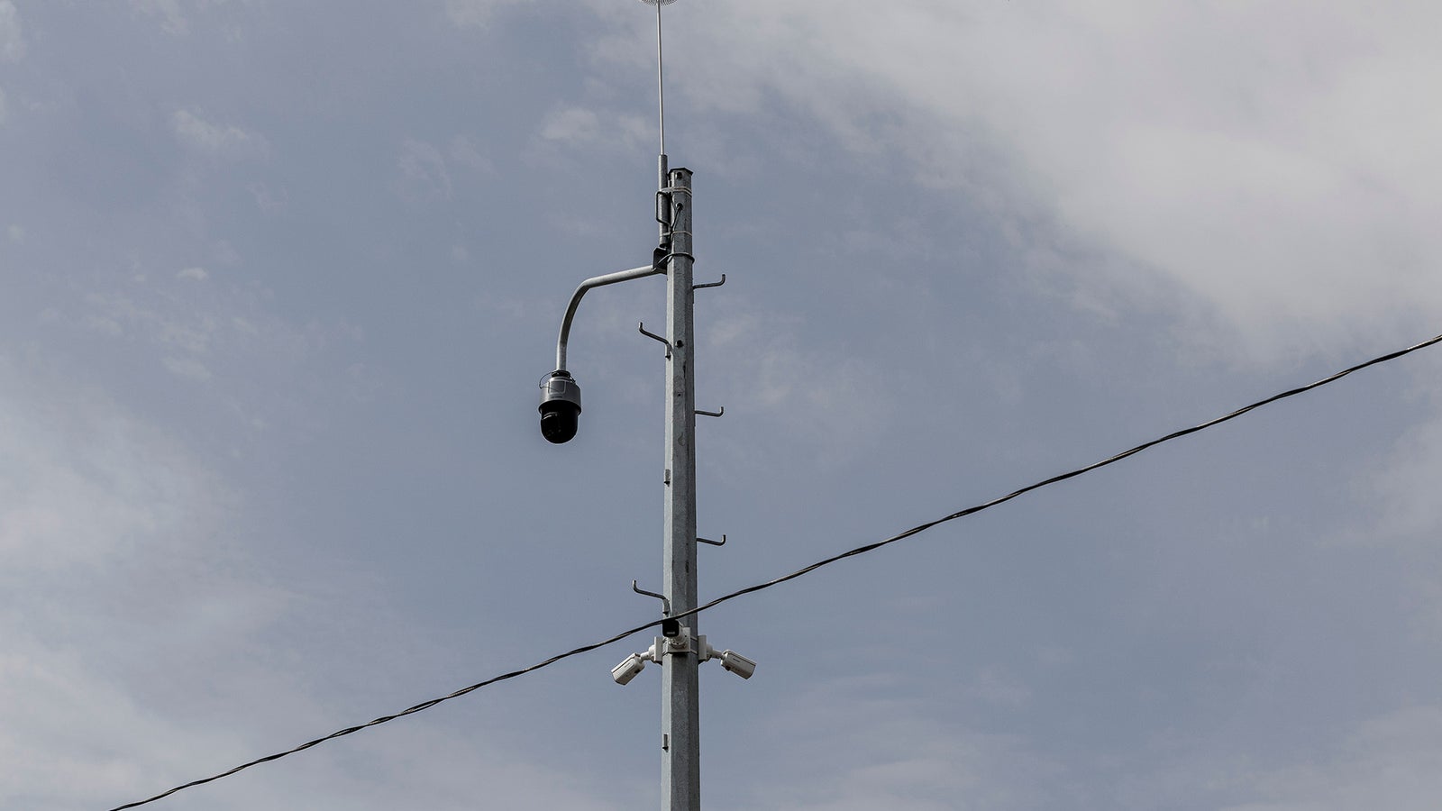 A tall metal pole with a security camera and several antennas against a cloudy sky, with power lines crossing in the foreground.