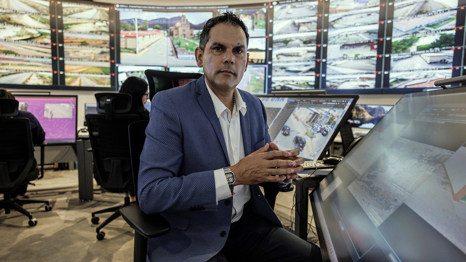 A man in a blue blazer sits in a tech control room surrounded by multiple large screens displaying surveillance footage.