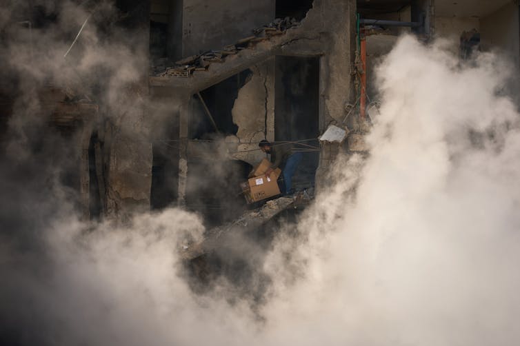 A man stands in a destroyed building as smoke rises around him.
