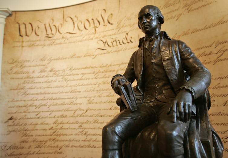 A stone statue of a man in front of a mural of the U.S. Constitution.