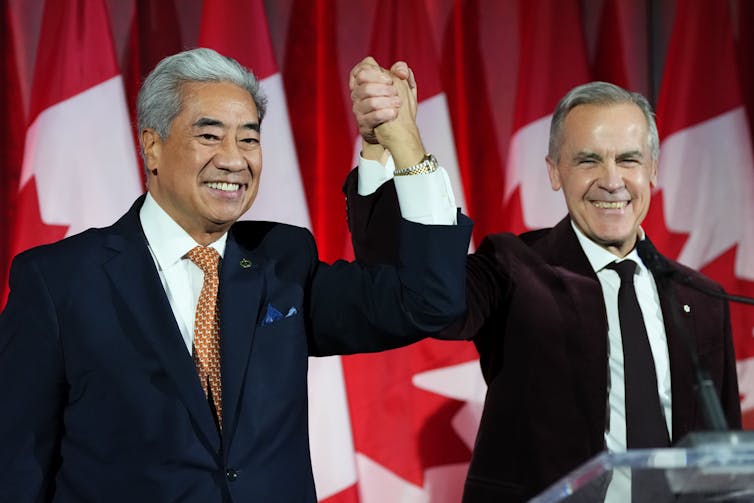 An Asian man and a man with short grey hair clasp hands and smile in front of a row of Canadian flags.