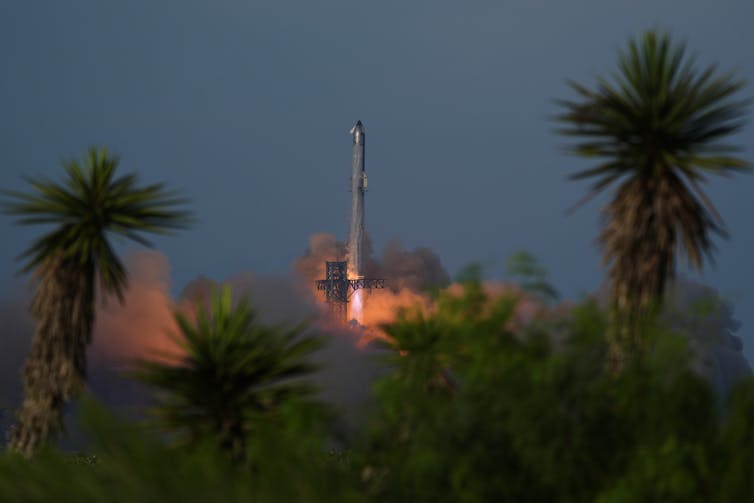 A rocket lifting off into the air, with a plume of smoke beneath it. In the foreground is a copse of trees.