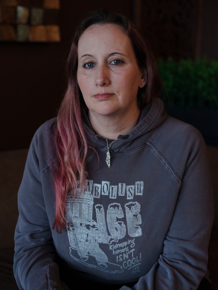 A woman with pink hair looks into the camera. She is wearing a sweatshirt that says “Abolish ICE, Kidnapping Humans Isn't Cool, It’s Evil.”