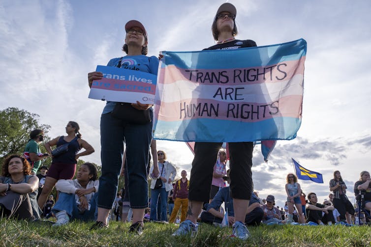 Two women standing in a field during a march holding up a trans flag in support of trans rights.