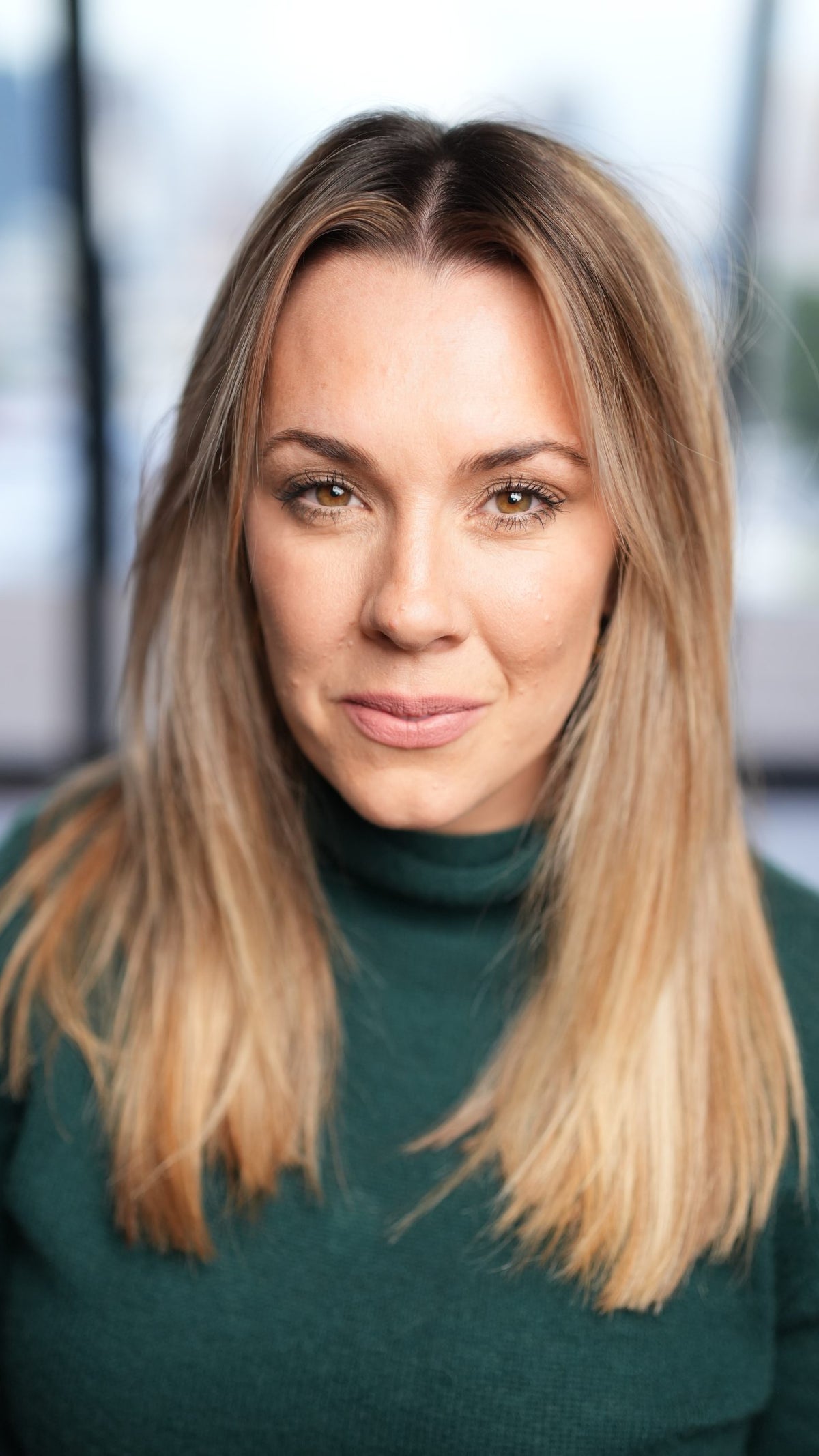 A woman with long, straight blonde hair and hazel eyes, wearing a green sweater, smiles at the camera indoors.