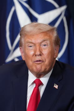 A man with fluffy white-blond hair and orange-ish skin with the NATO emblem behind his head.