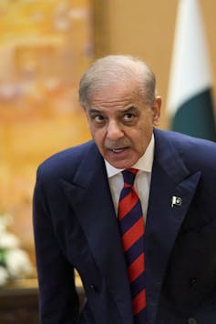 A man with grey hair wearing a blue suit and a blue and red striped tie sits down.