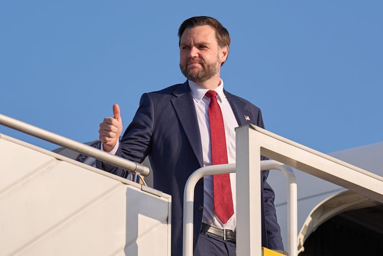 a man with dark hair and a beard gives a thumb's up as he boards an airplane
