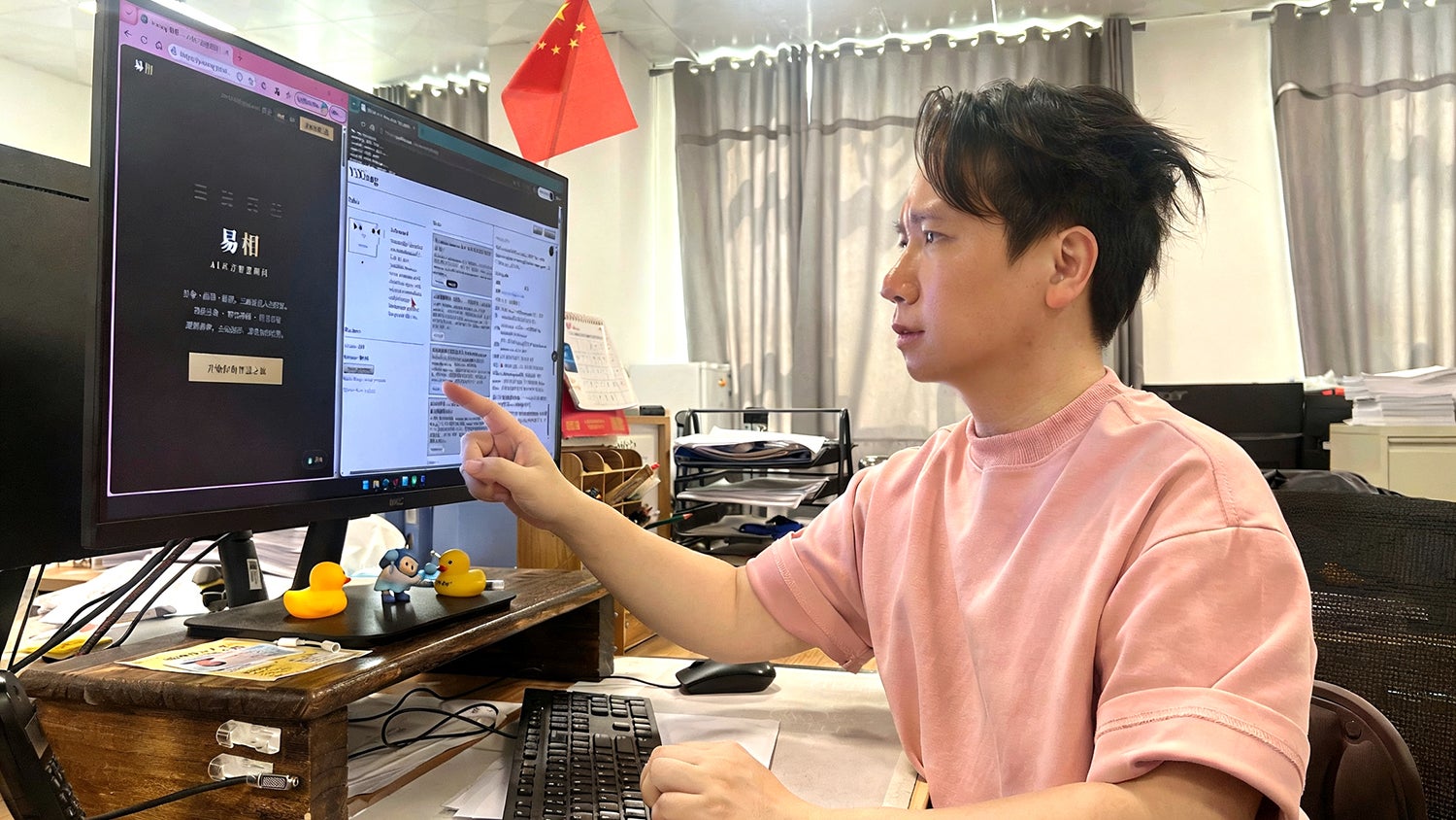 A person in a pink sweatshirt points at a computer screen displaying a website, with documents and a Chinese flag in the background.