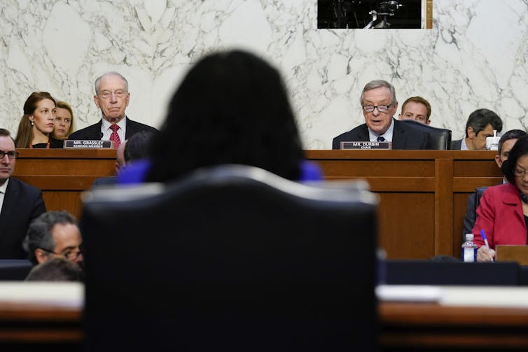 A Black woman with her back to the camera listens to a man speak behind a bench.