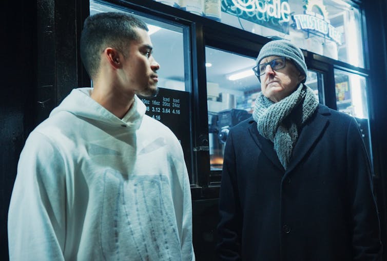 On a night street two men look at one another, warmly dressed.