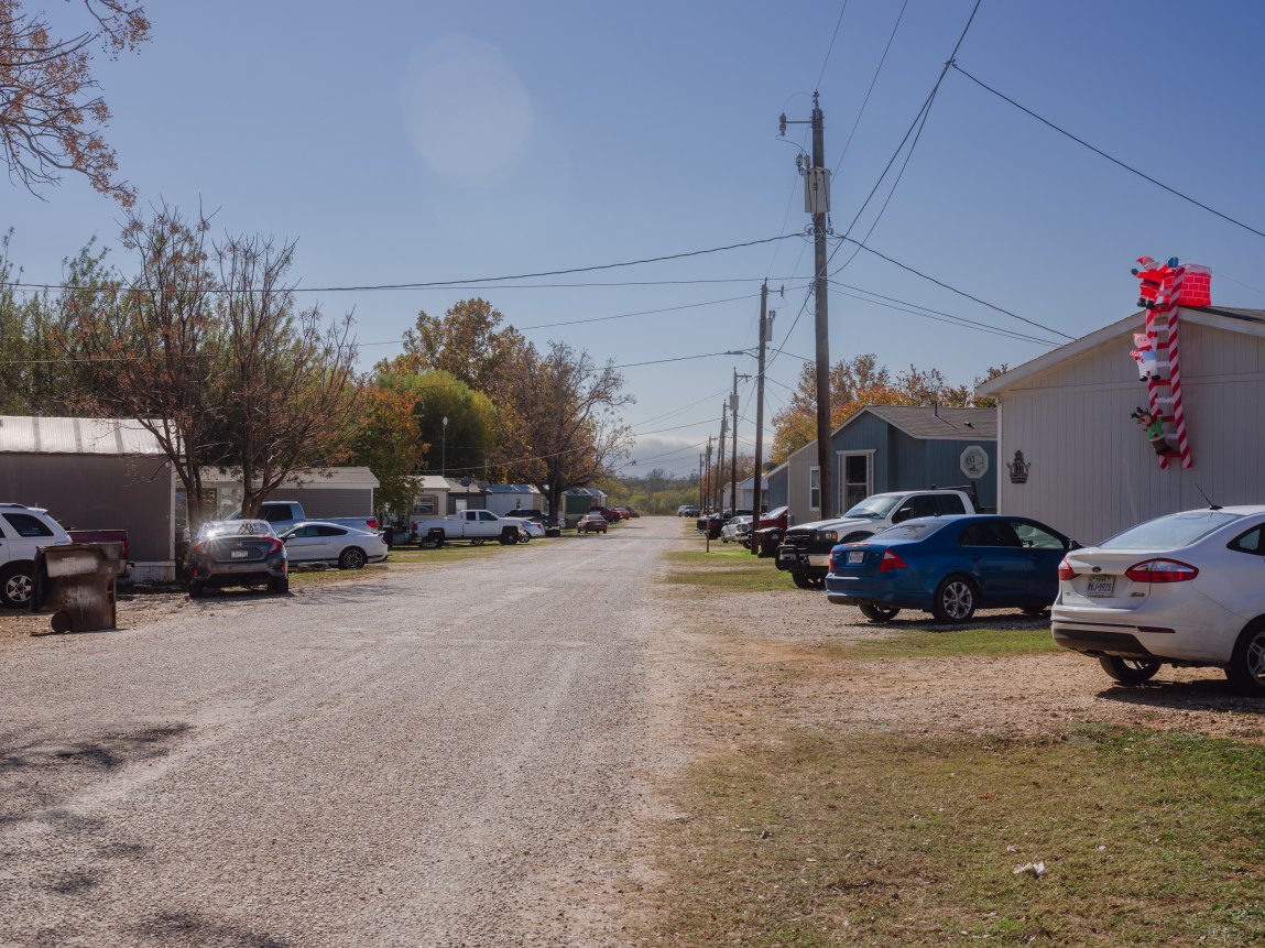 A gravel road runs between over a dozen cars parked alongside multiple single-wide housing units, with electricity wires running from a row of telephone poles.