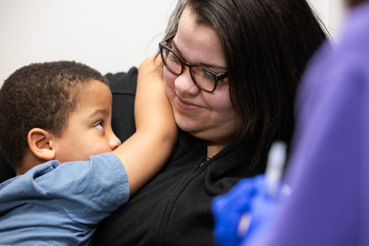 A small boy embraces and looks up at a woman smiling down at him. In the foreground, a blurry person wears blue gloves and a purple shirt.