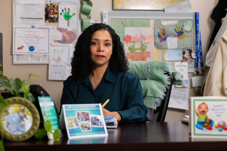 A woman sits at a desk with lead information flyers in front of her. Behind her, the wall is decorated with children’s drawings.