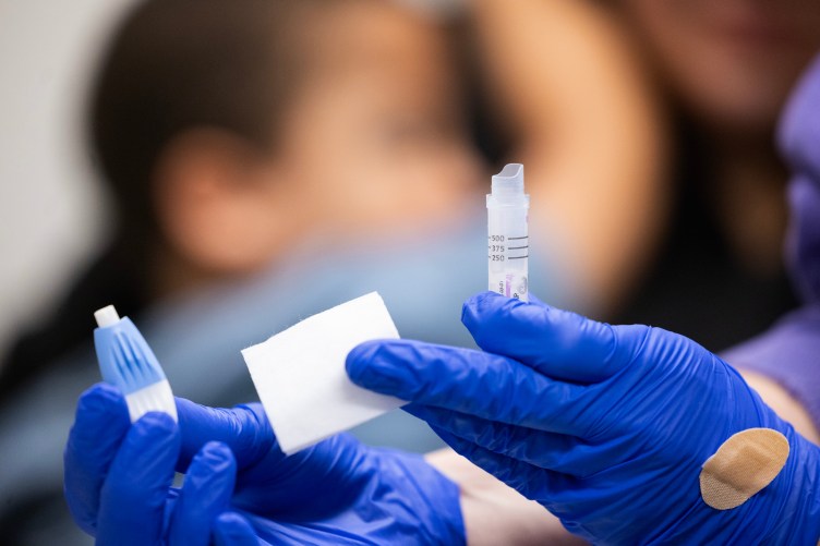 A close-up of someone’s hands in blue gloves holding medical supplies. Out of focus behind the hands is a child holding onto an adult.