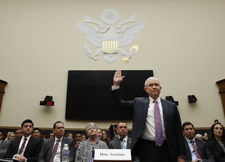 A man dressed in a suit and tie lifts his right hand in front of a panel of lawmakers.