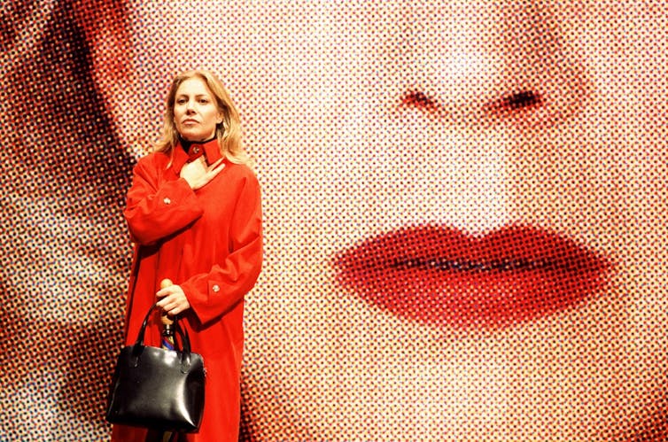 A woman in a long red coat, standing in front of a poster featuring a woman wearing red lipstick.