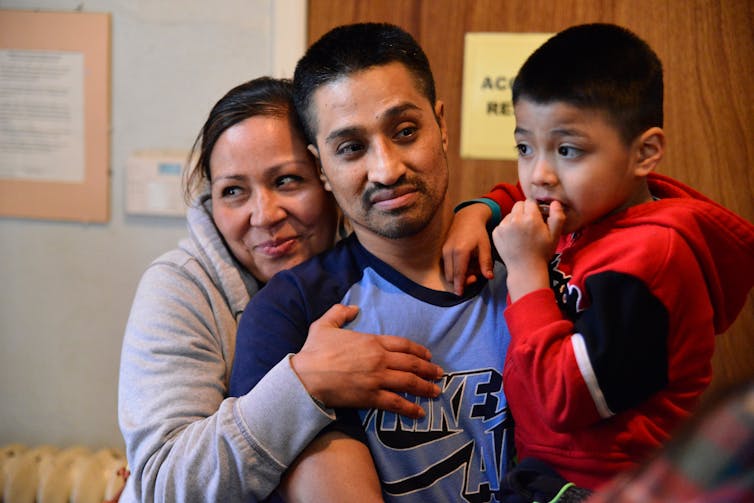 Man in blue shirt holds a child as woman hugs him from behind