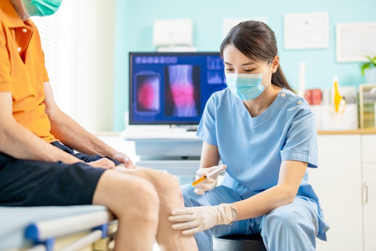 A female doctor or nurse wearing blue scrubs injects a needle into a patient's knee. The patient is seated on the examination table.