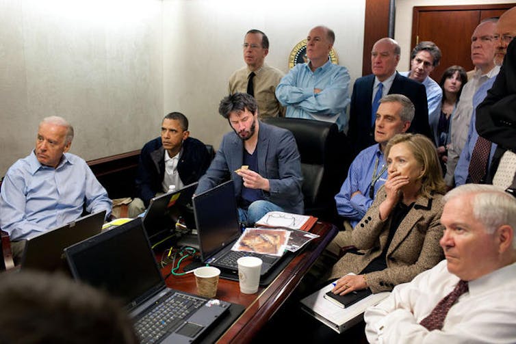 Keanu Reeves holding a sandwich, sitting at a conference table with laptops surrounded by U.S. leaders
