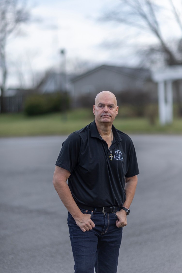A bald man wearing dark jeans, a polo shirt and a necklace with a cross stands with his hands in his pockets. The background is a blurred road and buildings with vegetation.