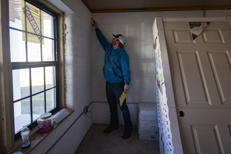 A man stands in a partially built house, pointing at a crack in one of the walls.