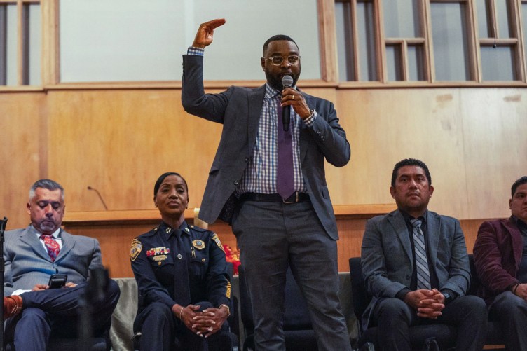 A man stands and speaks into a microphone while gesturing with his right hand. A woman in a police uniform and men in suits sit behind him.