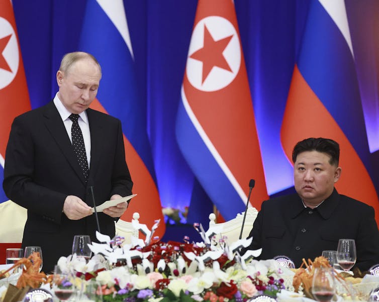 A balding white man speaks while looking at a document in his hand while an Asian man sitting down listens.