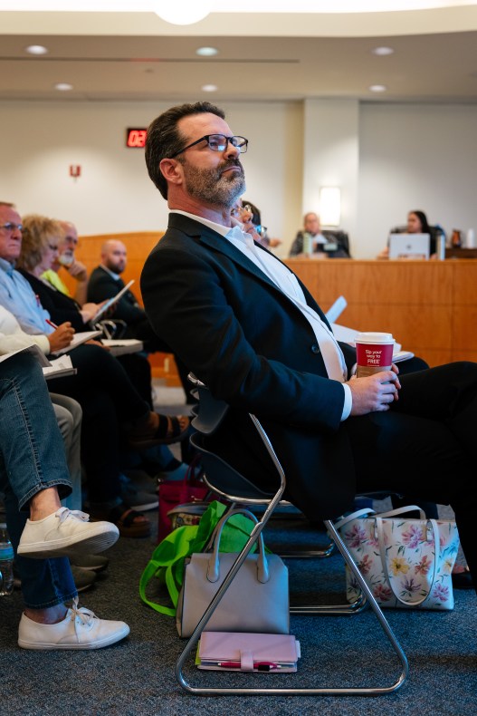 A man with dark hair and a short, graying beard, wearing a dark blue suit and a white shirt, sits in a chair during a meeting while holding a red, disposable coffee cup. Other meeting attendees sit in chairs behind him.