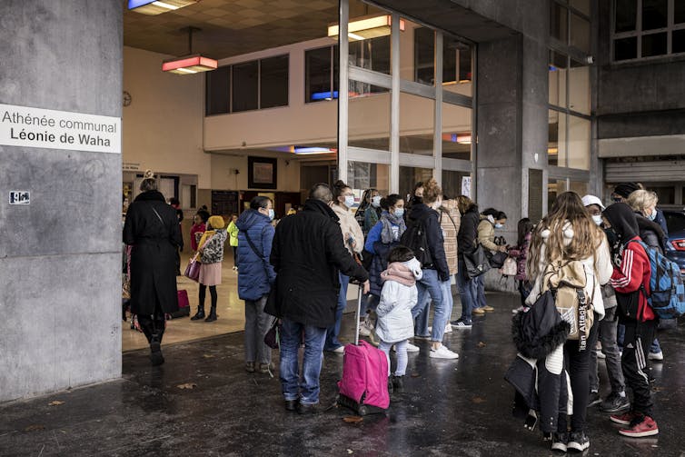 Young people in face masks congregating and chatting beside sliding doors in front of a concrete wall.