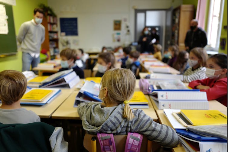 Children in face mask sit in desks in a square formation with teacher in a face mask at front.
