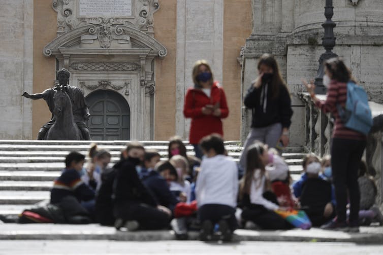 Young people with an adult seen outdoors, blurred against a statue in the background.