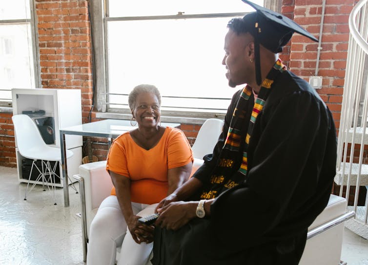 A young Black graduate in colourful stole and gown plus cap sits with an elderly Black woman with each smiling at the other.