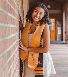 A young woman in a formal dress and colourful African stole and also a bronze stole stands leaning against a wall looking proud and happy.