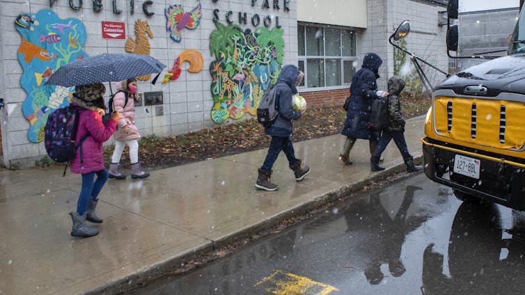 Students with umbrellas seen on a sidewalk near a school bus.