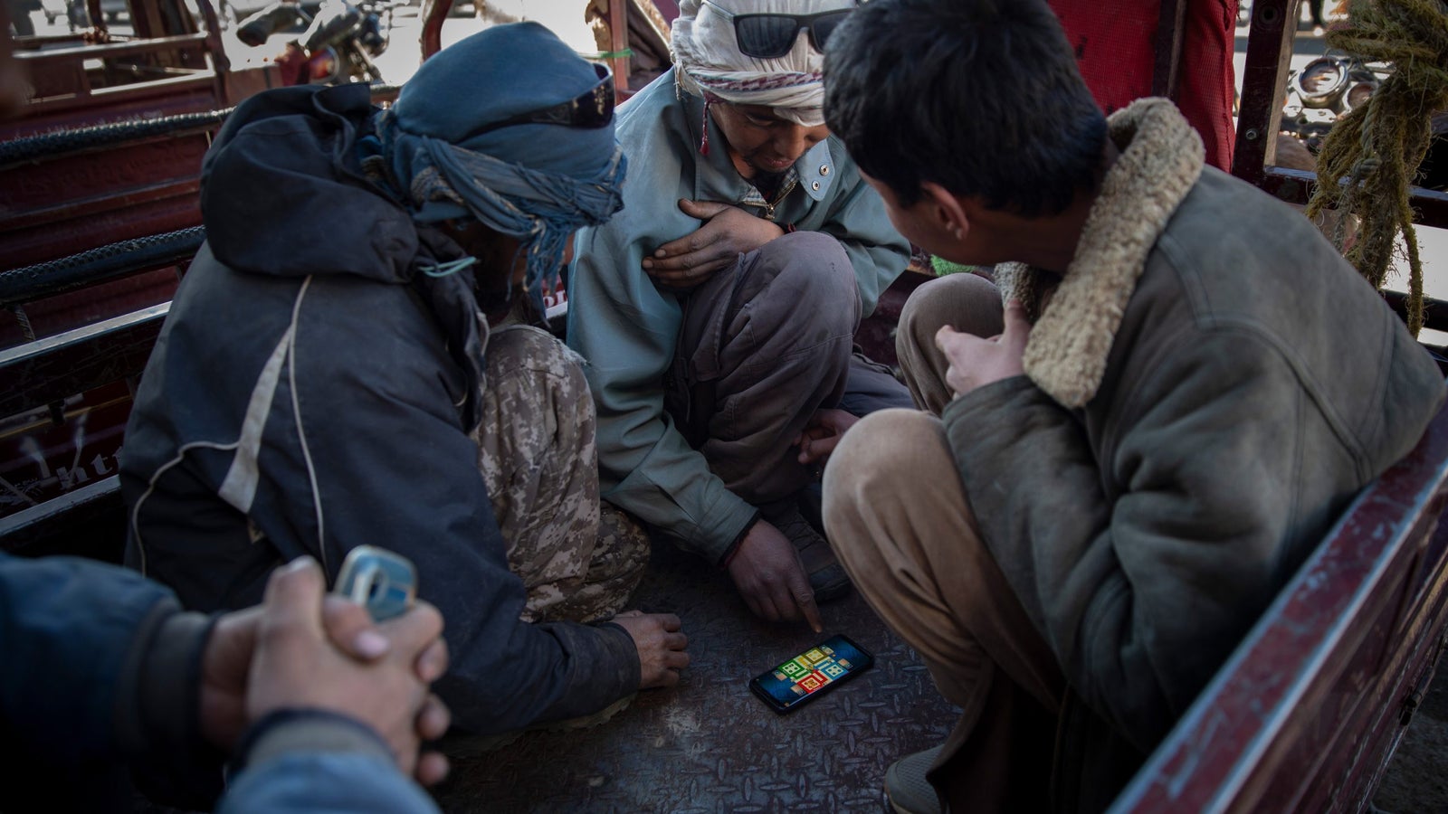 Three men sit in a truck bed, intently focused on a smartphone displaying a game or app, with various backgrounds visible.