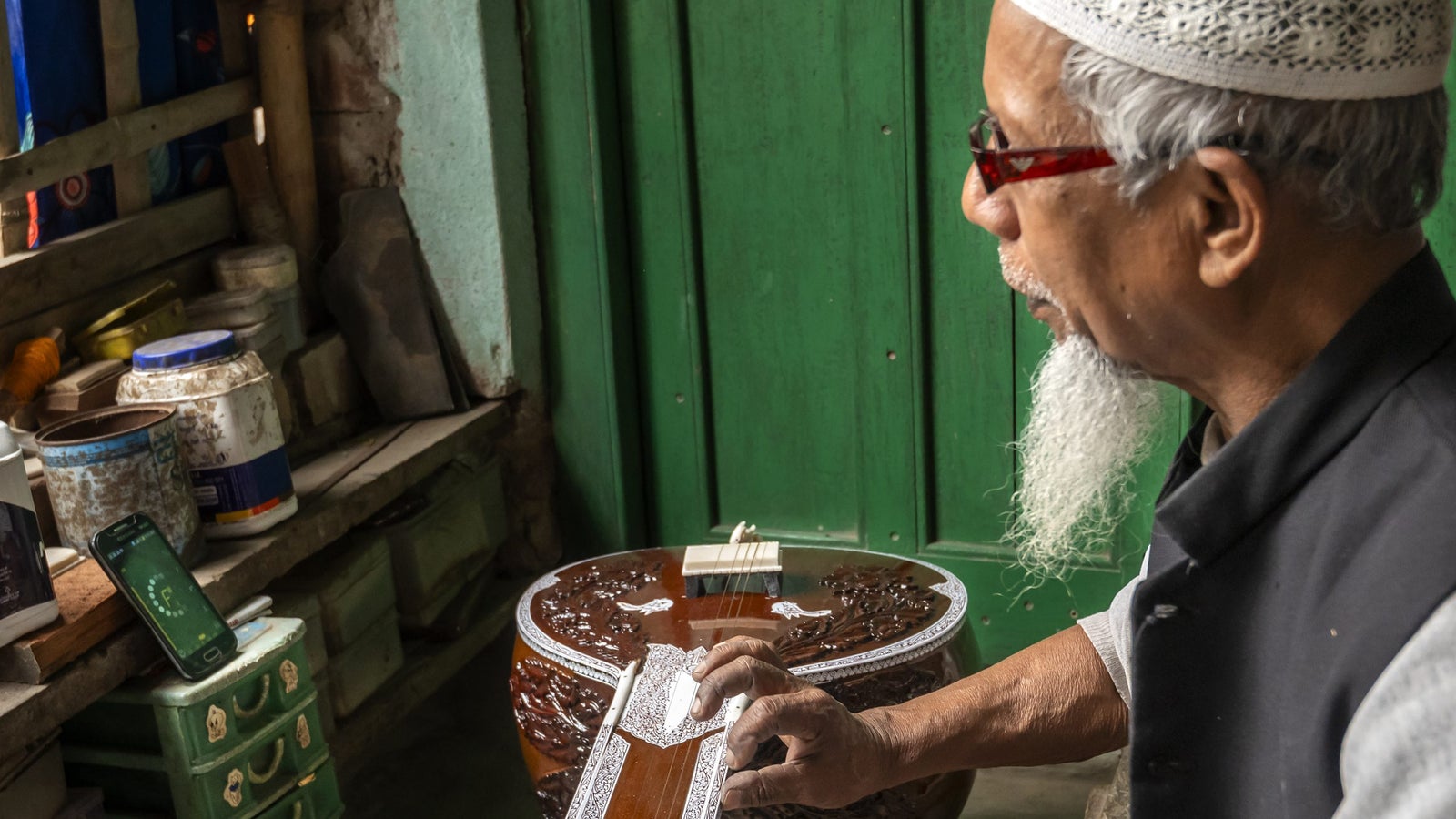An elderly man with glasses and a white cap focuses on a wooden musical instrument while a phone displays information nearby.
