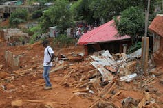 A man stands on top of thick red mud next to broken homes that have been damaged by a landslide